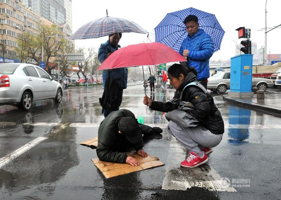 4月16日，山东青岛，清晨7时许，一名老人在威海路红路灯路口因下雨路滑摔倒，卧在积满雨水的道路上，几次试图爬起来都没能成功。旁边走过来一名撑伞的中年妇女，弯腰与老人交谈，此处是红绿灯路口，不少机动车从他们身边驶过。中年妇女蹲下身，将雨伞伸到老人上方，为其遮雨，全然不顾自己被淋湿。考虑到老人摔倒时有可能会出现骨折，贸然扶起容易加重伤情，市民们在为其遮雨挡车的同时，赶紧拨打120求助。
