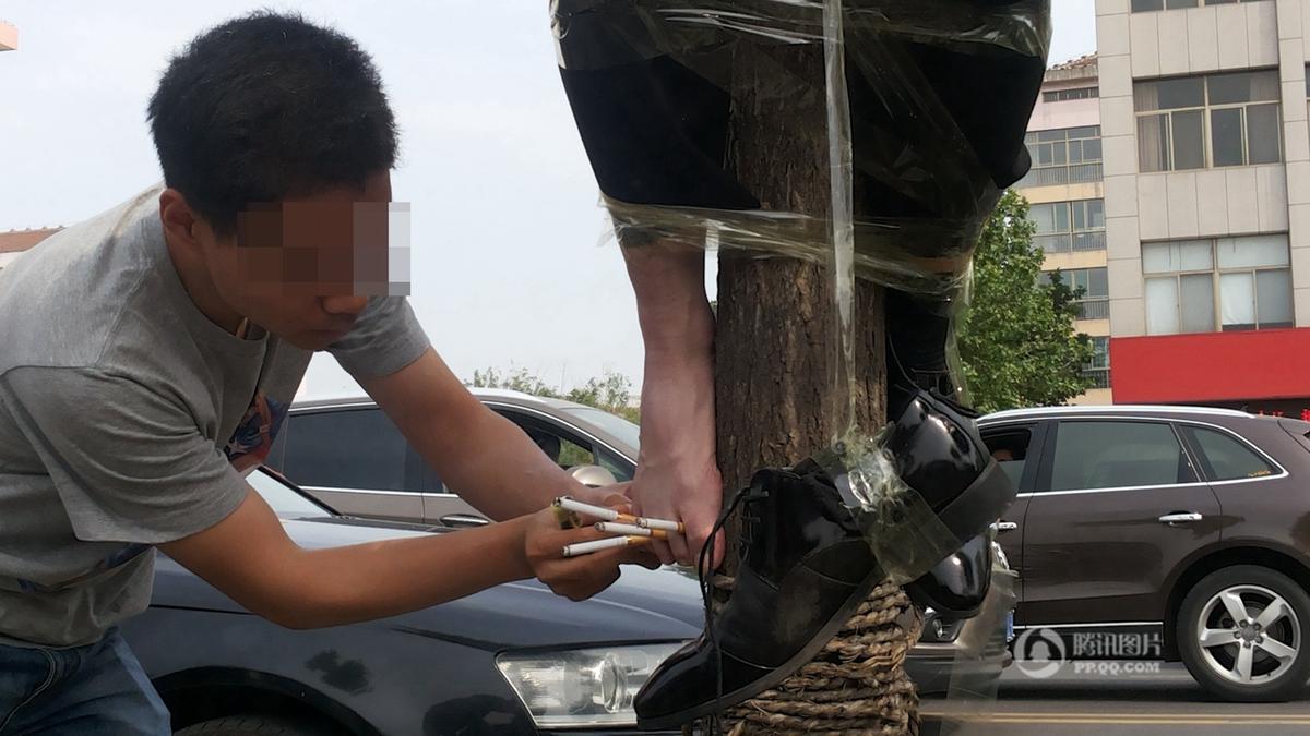 　　新郎被人用胶带捆绑在马路旁的大树上，一人正在其脚上点烟。